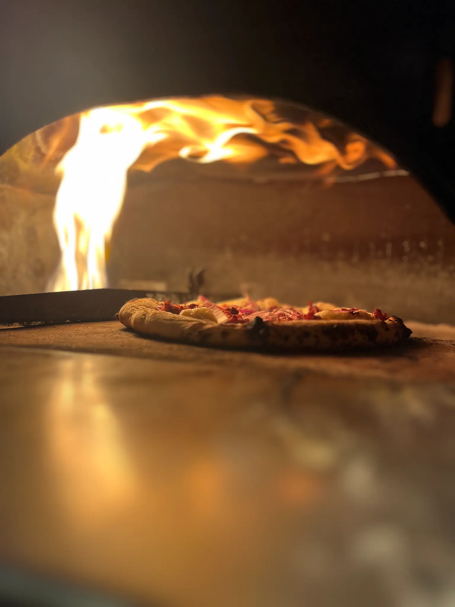 Pizza assando sobre a base do forno, com chamas acesas acima e a borda da pizza em primeiro plano.
