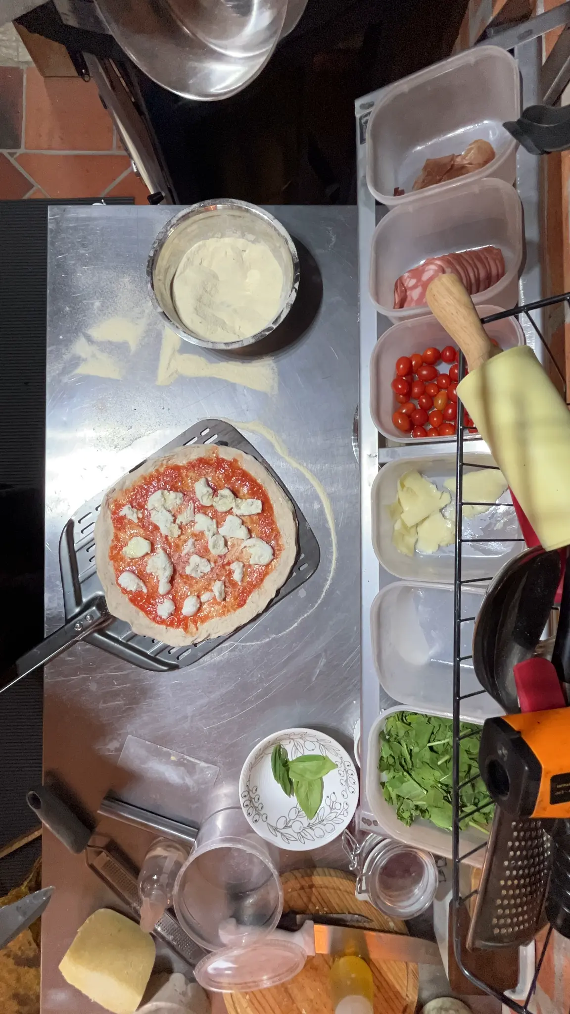 Vista de cima da bancada de preparo com massa aberta, molho e porções de ingredientes; ao redor há embutidos fatiados, tomates-cereja, queijo e folhas verdes.