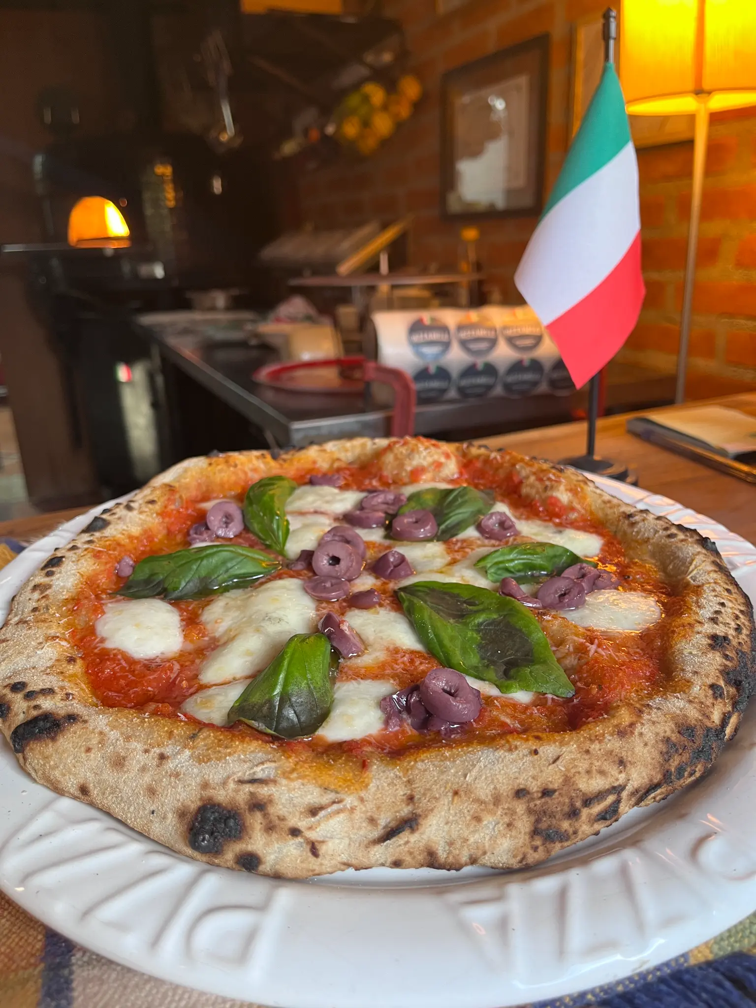Pizza redonda em um prato branco, com molho vermelho, queijo branco, folhas verdes e azeitonas roxas; uma bandeira da Itália aparece ao fundo.