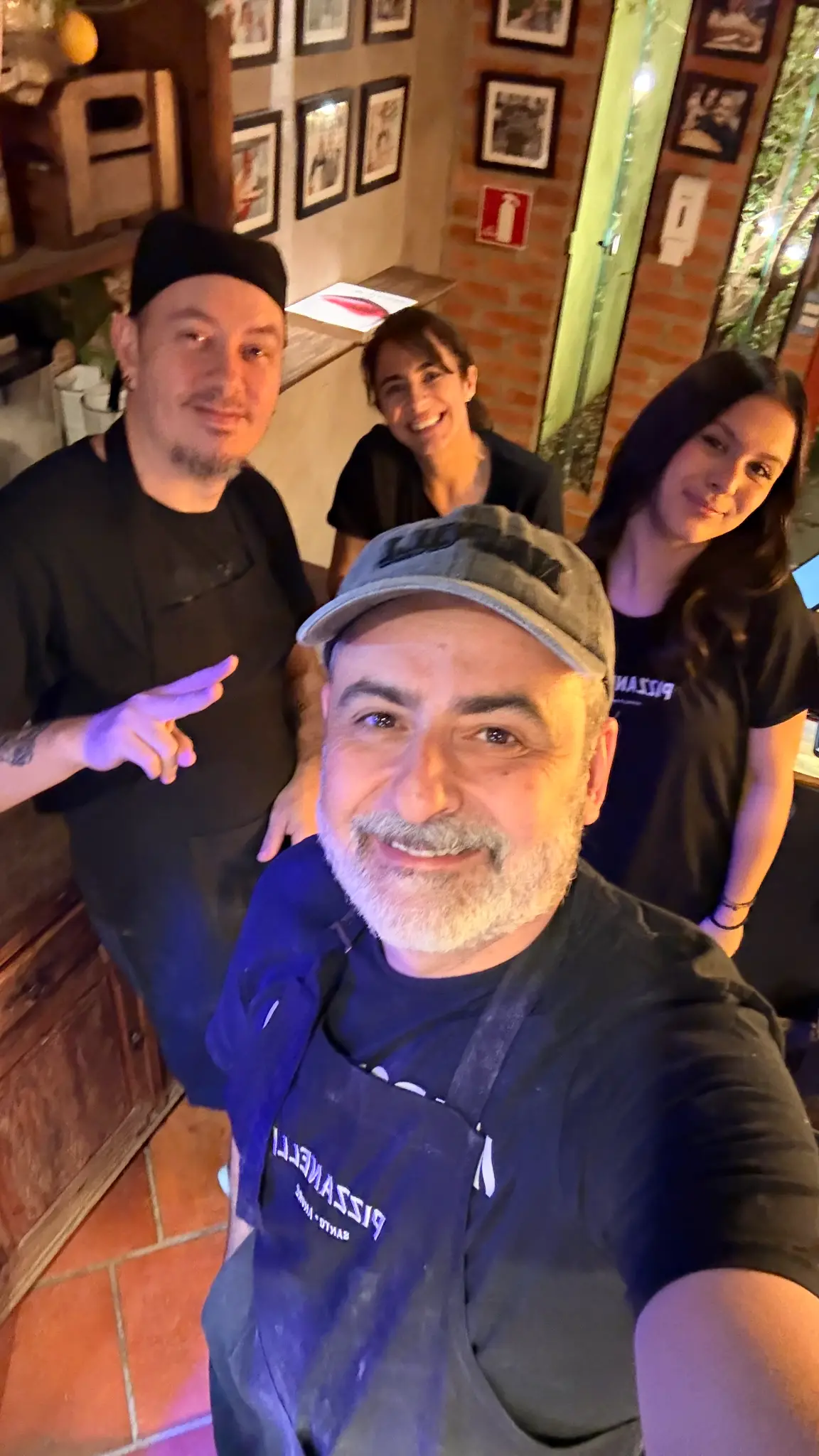 Selfie de quatro pessoas no interior da Pizzanelli, com balcão, quadros e uma parede de tijolos ao fundo.