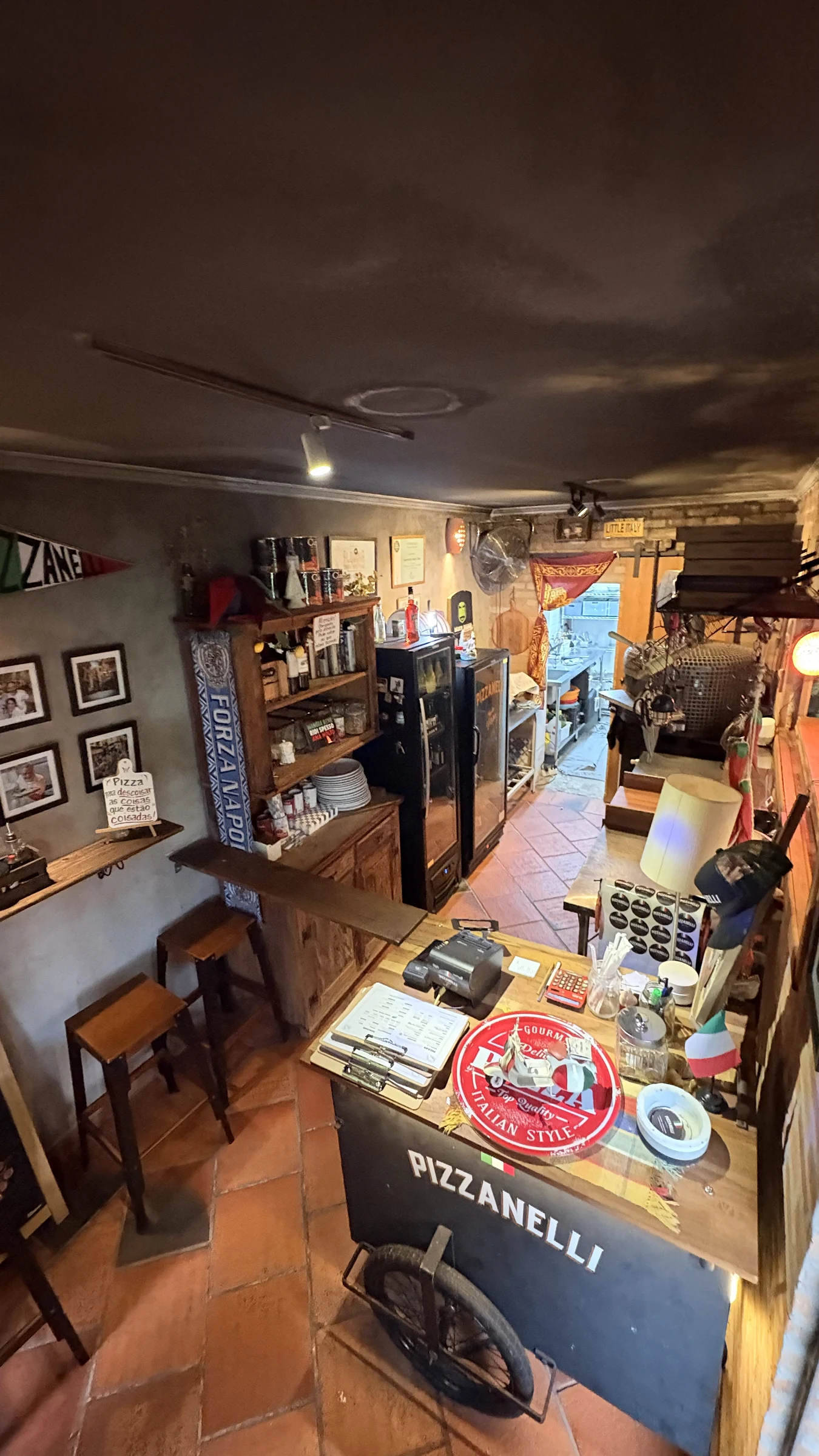 Interior da Pizzanelli com balcão de atendimento, bancos, prateleiras, quadros, uma bandeira italiana e o forno ao fundo.