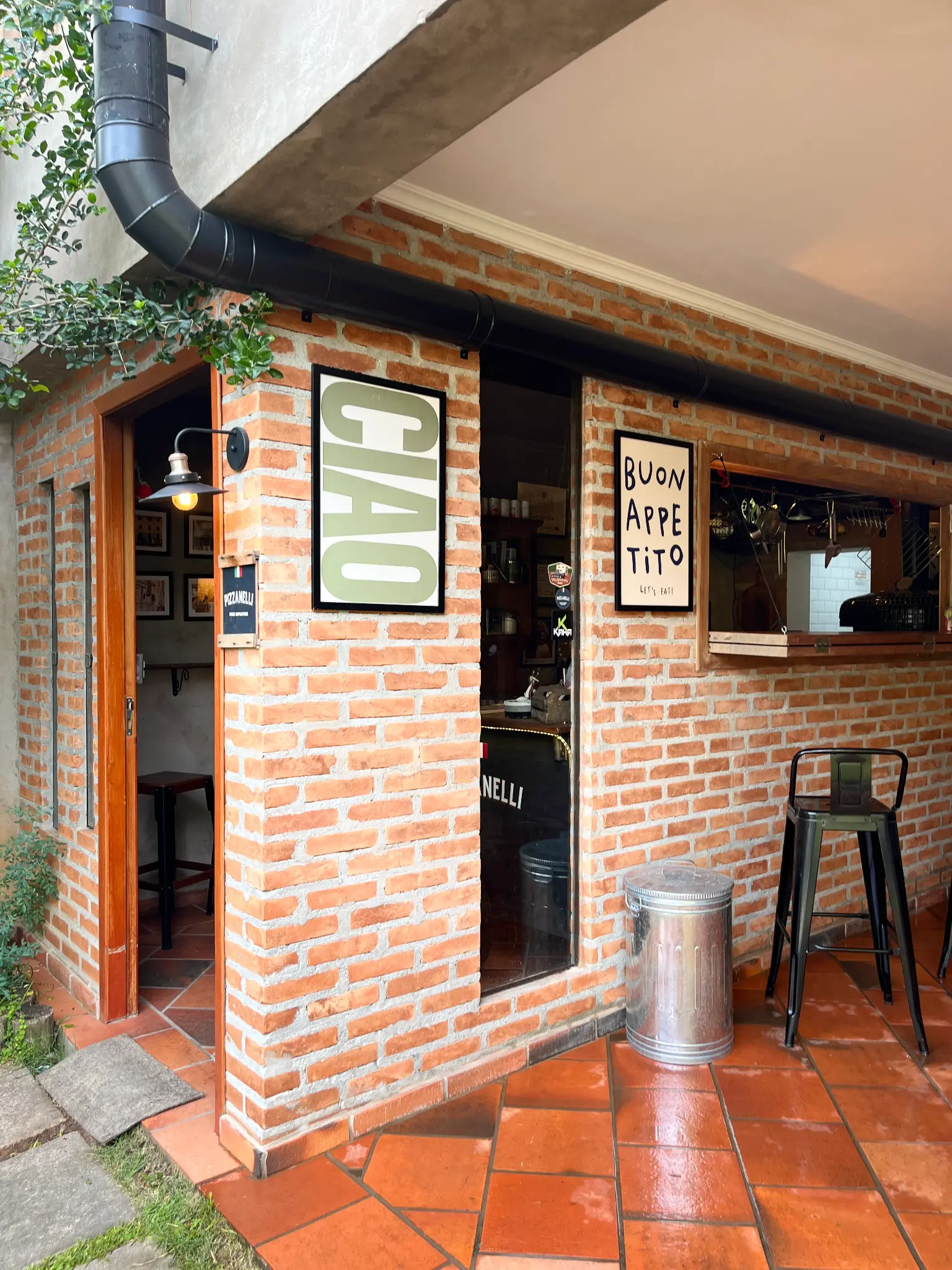 Fachada de tijolos da Pizzanelli, com porta aberta, janela de atendimento, placas decorativas e bancos altos na área externa.