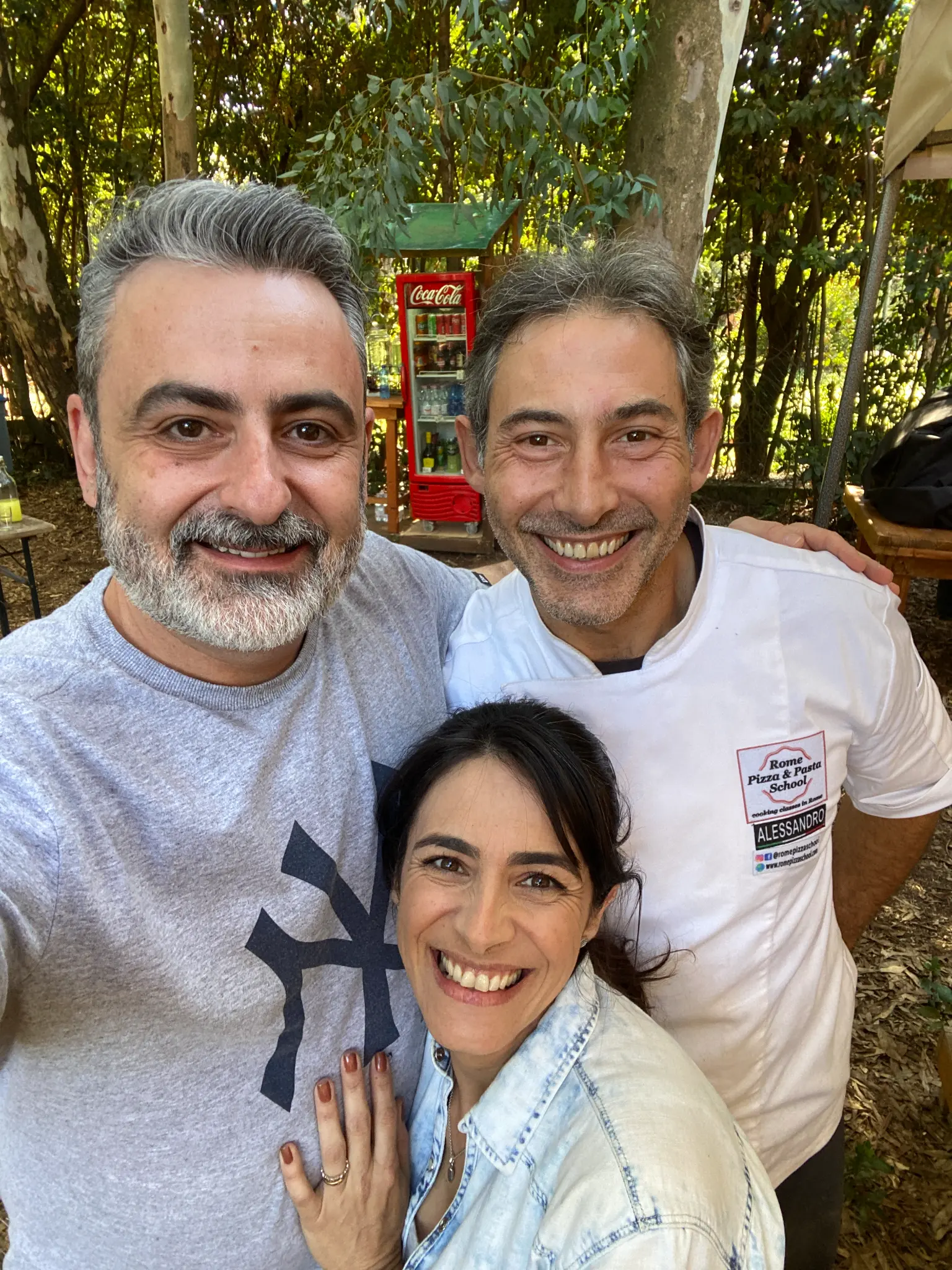 Três pessoas sorriem em uma área arborizada; o Maestro Alessandro D’Ascenzi usa a camiseta da Rome Pizza & Pasta School.