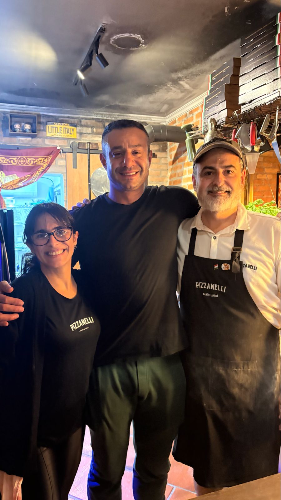 Três pessoas sorrindo lado a lado dentro da Pizzanelli, diante do balcão e dos equipamentos da casa.