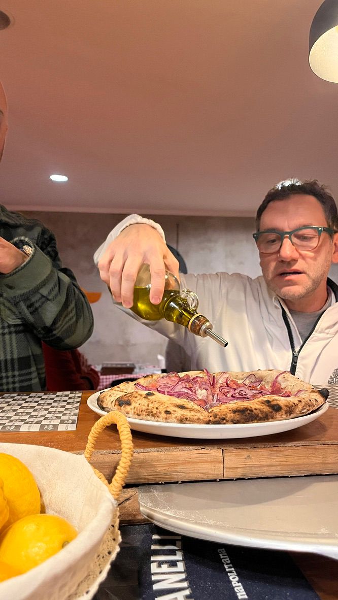 Pizzaiolo de óculos finaliza uma pizza no prato, regando-a com azeite antes de servir.