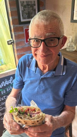 Homem de óculos segura um sanduíche recheado servido em um prato, dentro da pizzaria.