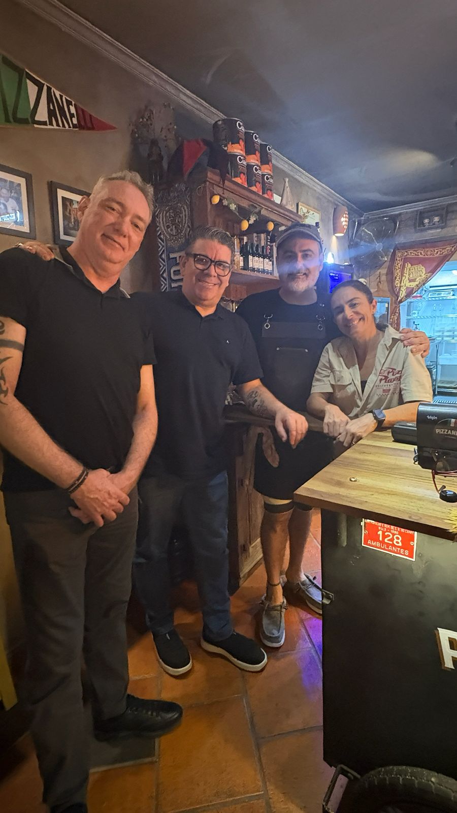 Quatro homens posam no salão da Pizzanelli, junto ao balcão e a uma mesa da casa.