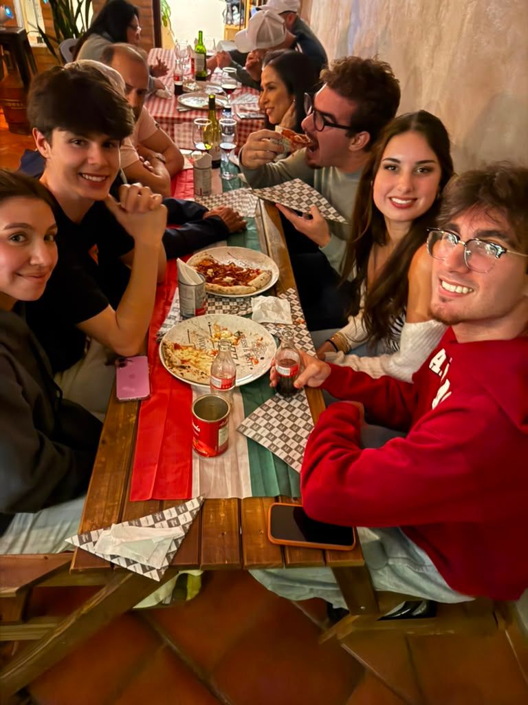 Grupo de amigos reunido em torno de uma mesa com pizzas, pratos e bebidas.
