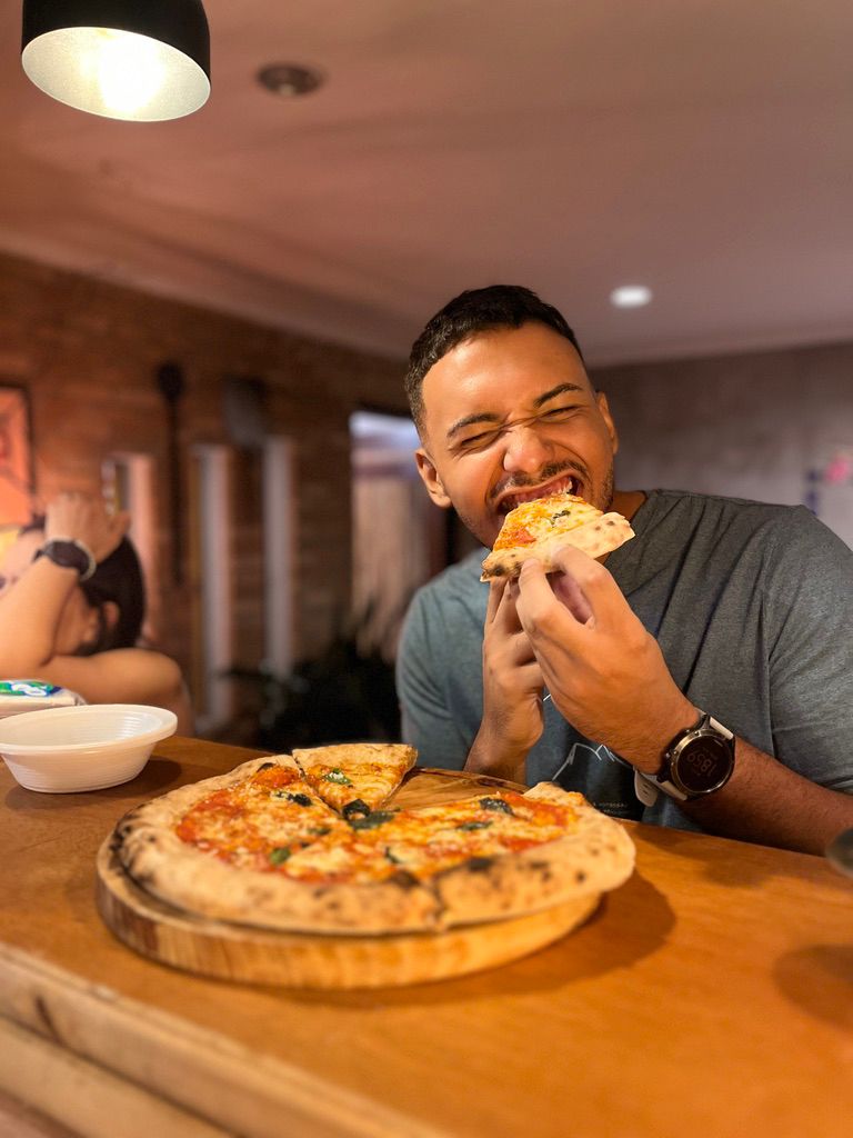 Homem prova uma fatia de pizza em uma mesa da Pizzanelli, com outra pizza servida à frente.