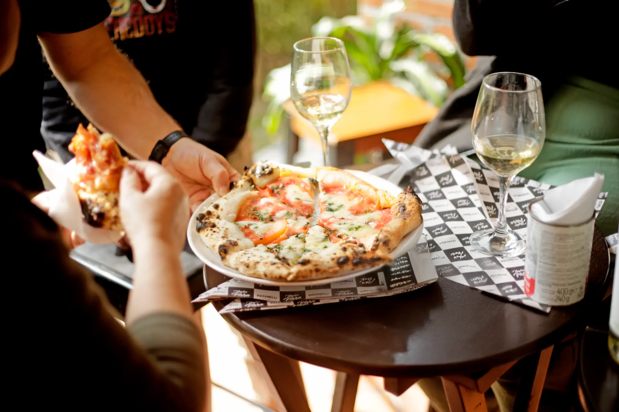 Pizza redonda em prato branco sobre uma mesa, com cobertura clara, áreas vermelhas e folhas verdes; uma pessoa segura uma fatia à esquerda e há duas taças ao fundo.