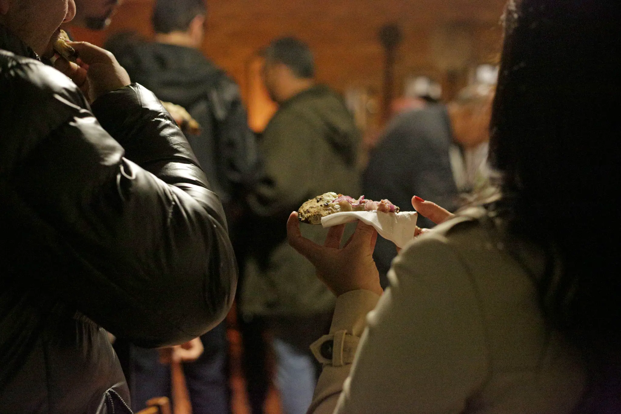 Pessoa segura um pedaço de pizza com elementos claros, verdes e roxos, parcialmente envolto em guardanapo, enquanto outras pessoas aparecem ao redor desfocadas.