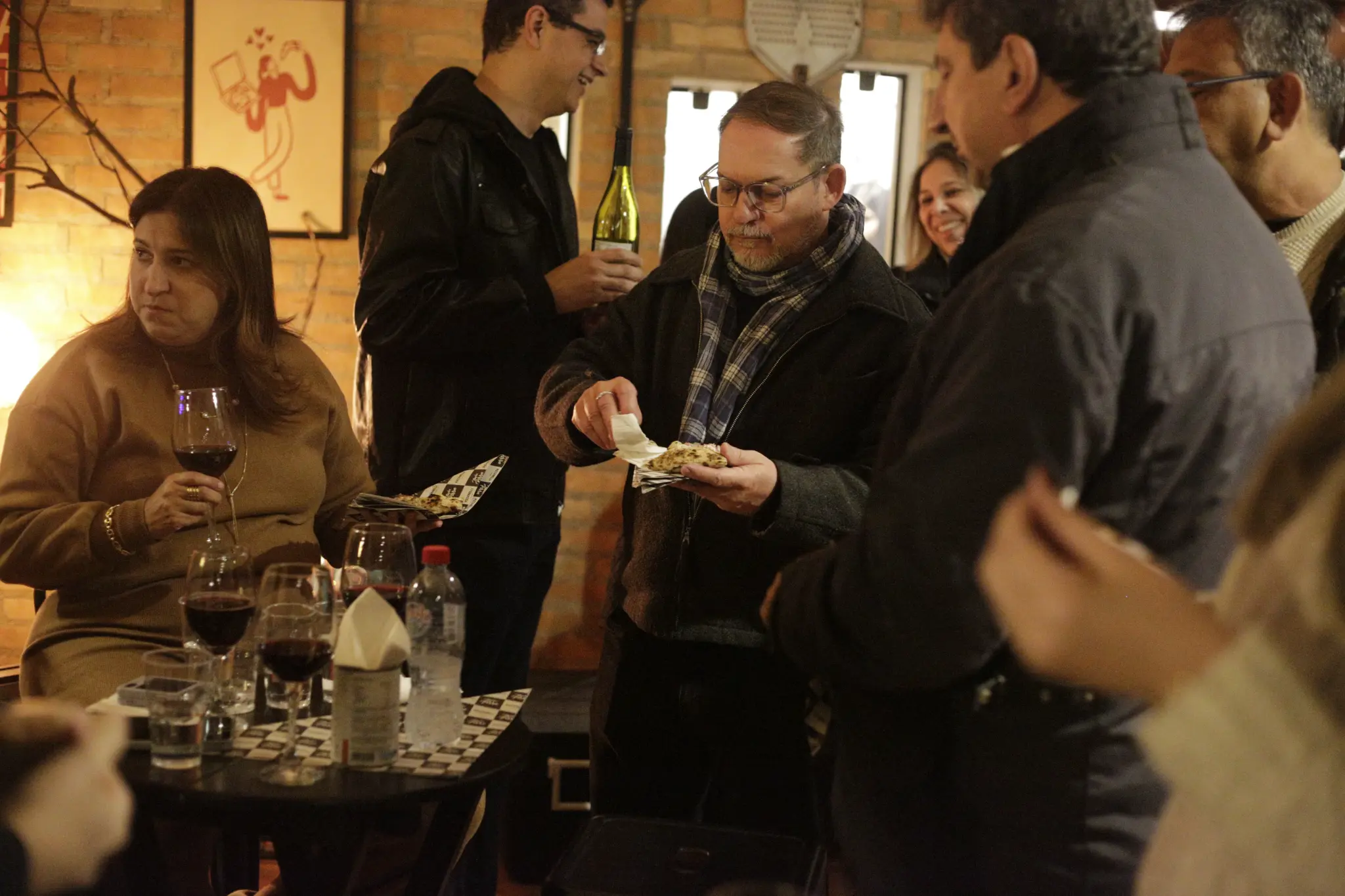 Grupo reunido no salão; uma pessoa ao centro segura um pedaço de pizza e um guardanapo, enquanto há taças e copos nas mesas.