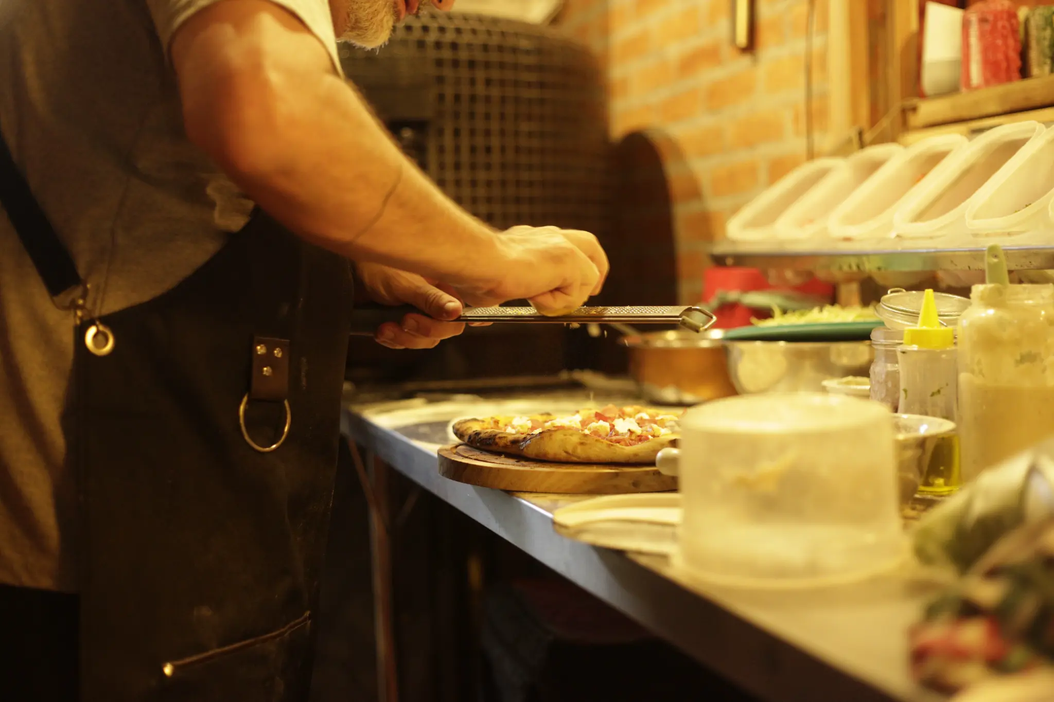 Pessoa usando avental rala queijo sobre uma pizza em uma bancada de cozinha; recipientes de ingredientes e uma parede de tijolos aparecem ao fundo.