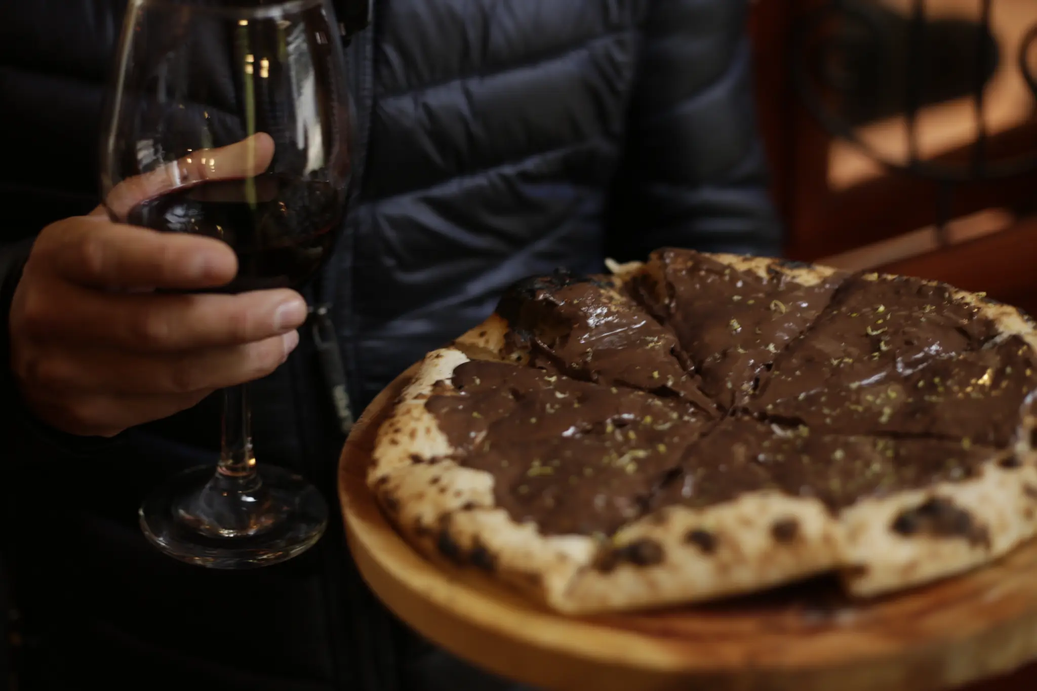 Pizza cortada em fatias, com cobertura escura e brilhante, servida em uma tábua de madeira ao lado de uma taça com bebida escura.