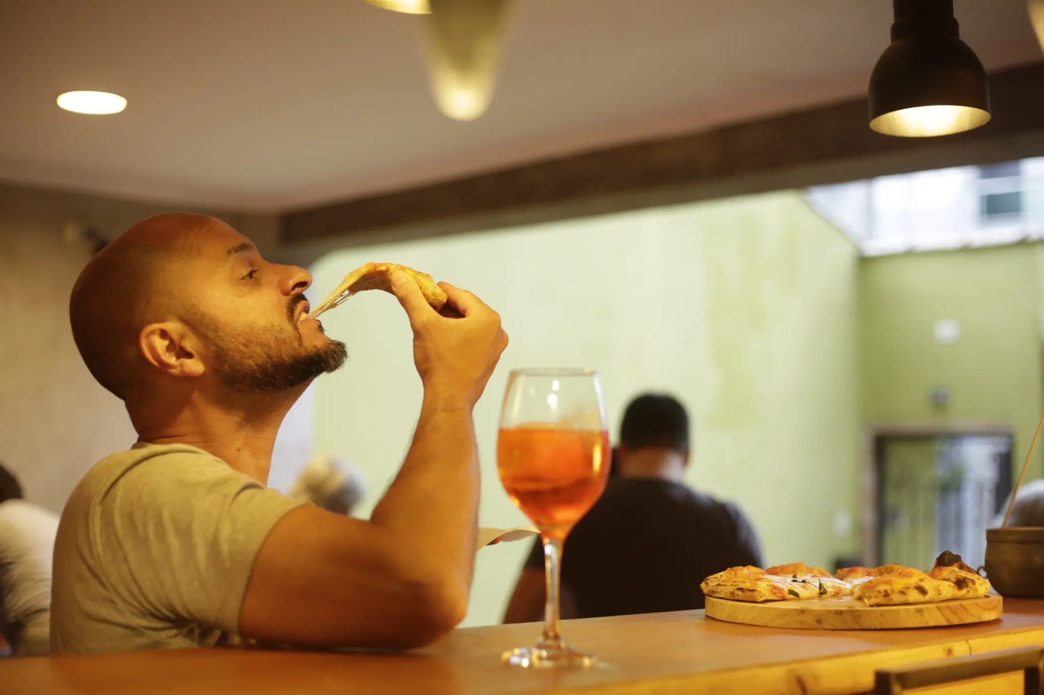Pessoa sentada à mesa leva uma fatia de pizza à boca, com queijo esticado; à frente há uma taça com bebida alaranjada e uma pizza sobre tábua de madeira.