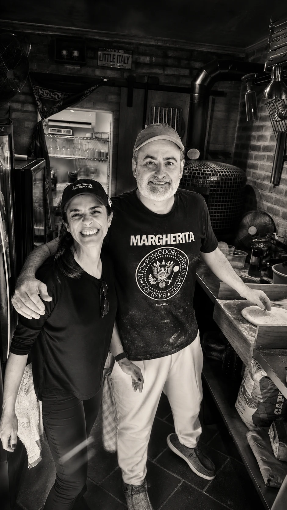 Duas pessoas sorriem e posam juntas em uma cozinha de pizzaria; uma veste camiseta com a palavra MARGHERITA e equipamentos aparecem ao fundo.