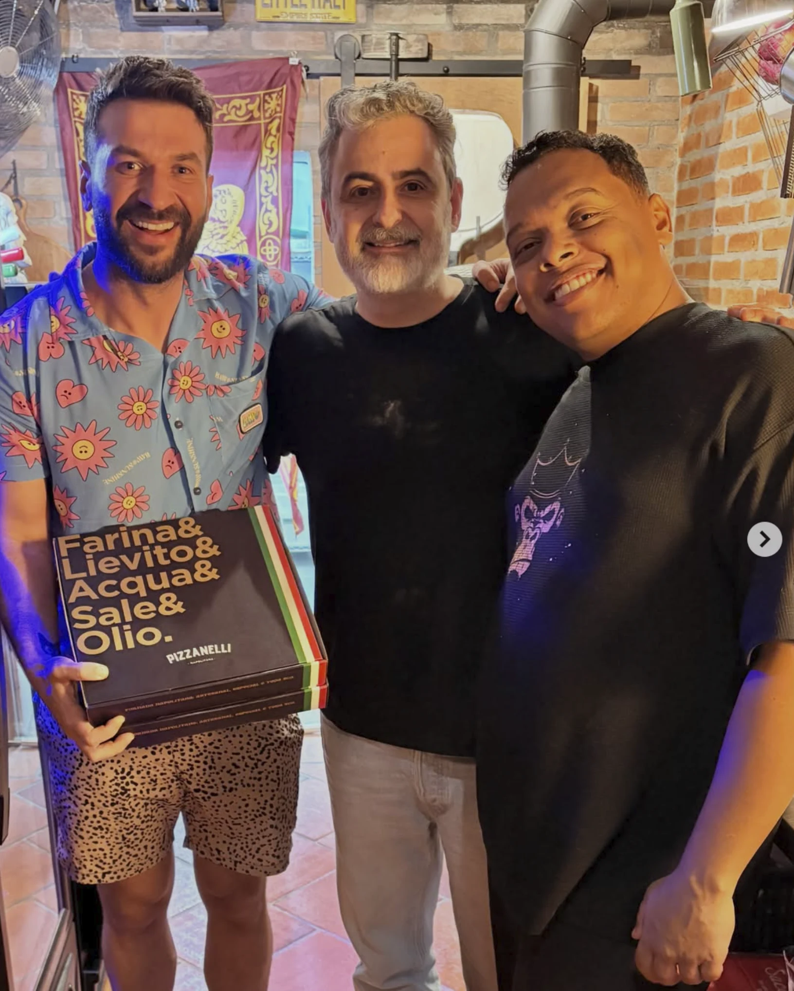 Três pessoas sorrindo no interior da Pizzanelli; uma delas segura caixas da casa, com parede de tijolos ao fundo.