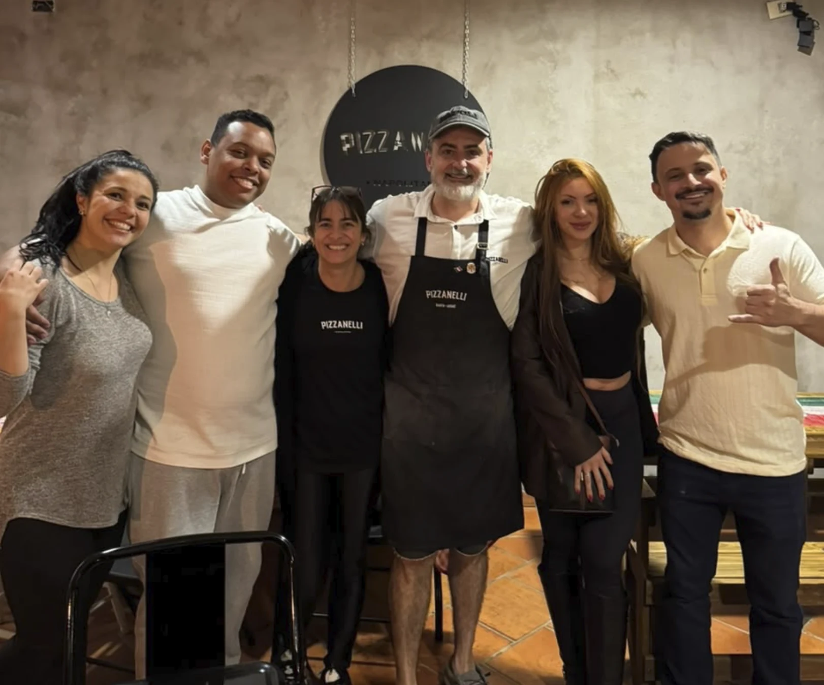 Seis pessoas posam juntas no salão, diante do logo da Pizzanelli; duas usam peças com a marca da casa.