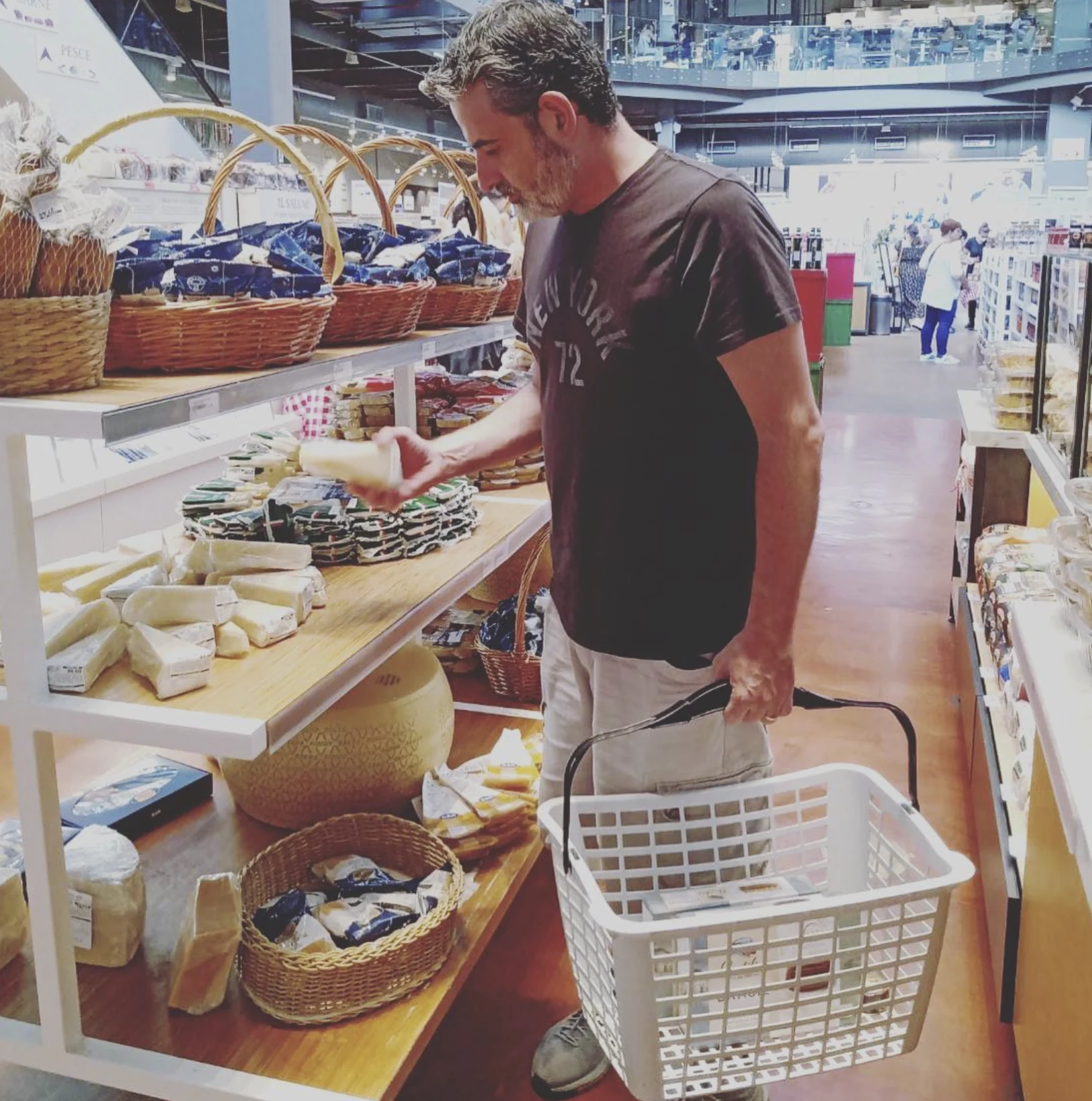 Pessoa observa uma embalagem na seção de queijos de um mercado, com uma cesta de compras na outra mão.