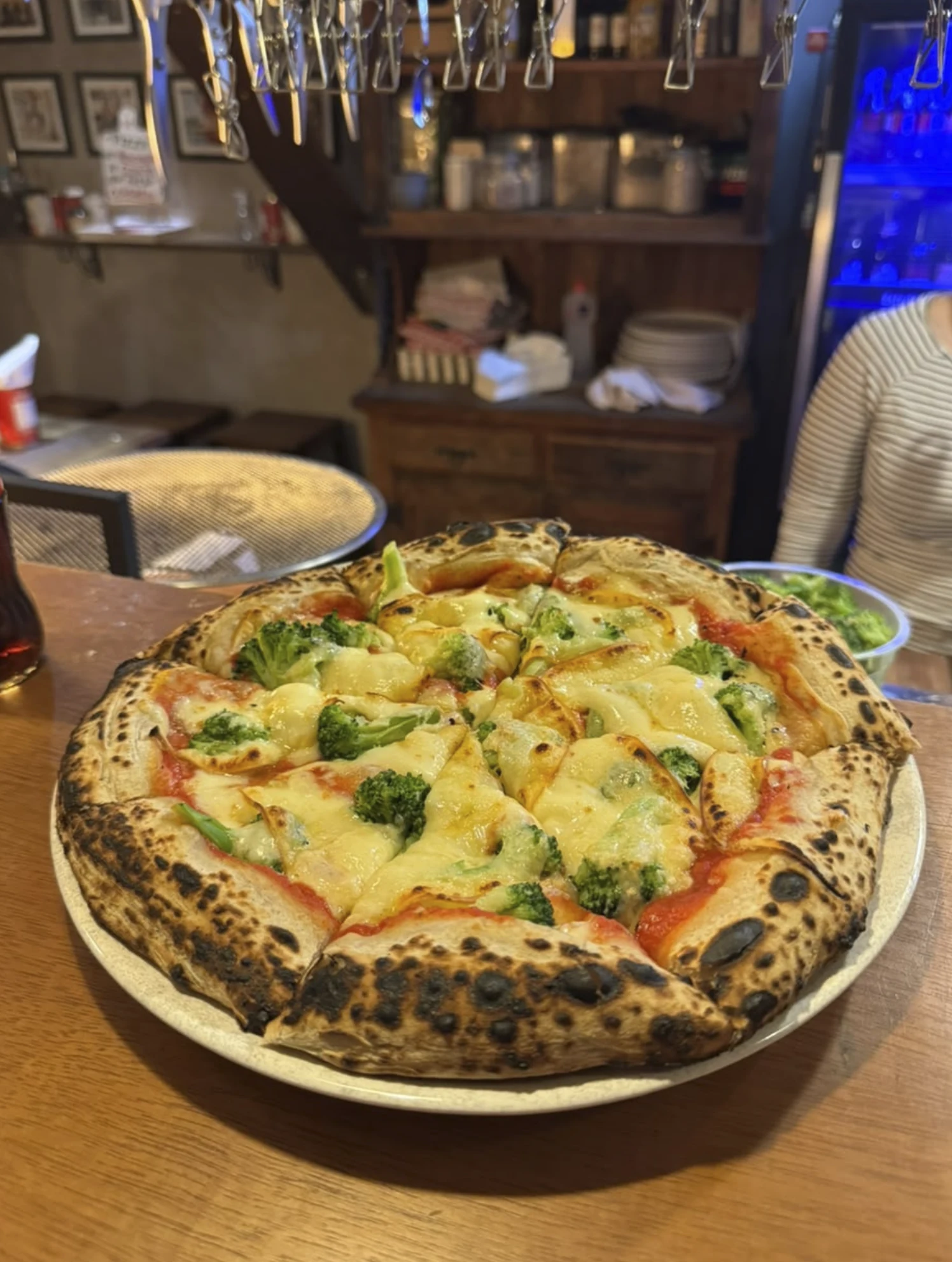 Pizza redonda em prato branco, com molho de tomate, queijo derretido e floretes verdes; o salão aparece ao fundo.